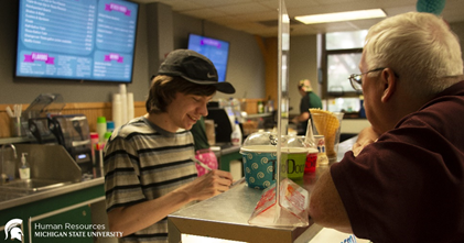 Spartan Project SEARCH graduate Cash helping a male customer at the counter in the MSU Dairy Store.